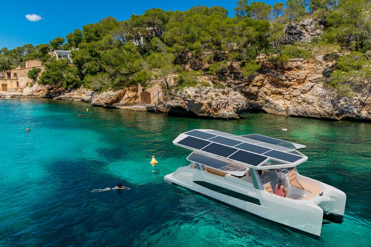 Millikan M10 in clear blue water and green forest in background on rocky cliffs. The boat has a solar panel roof