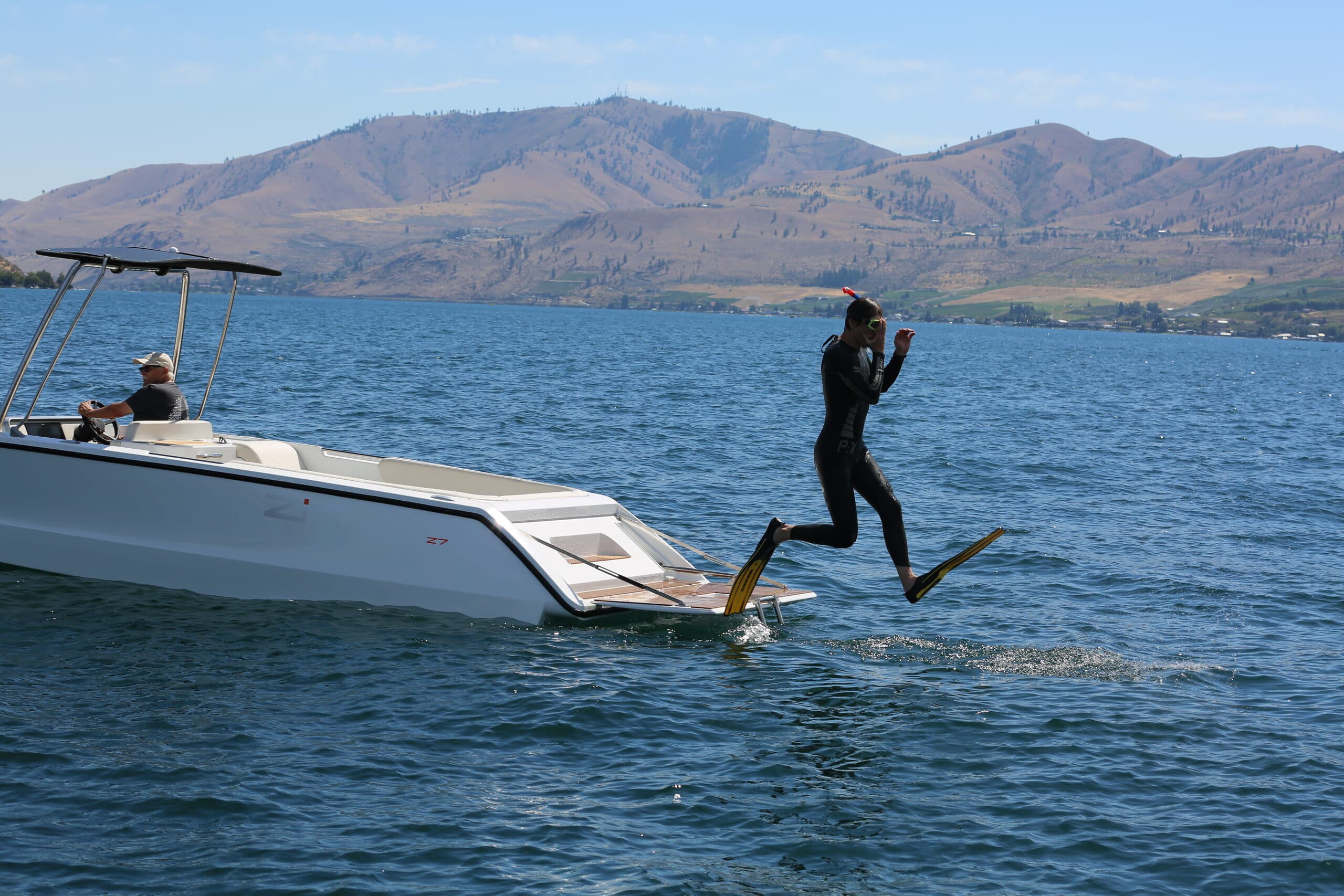 Scuba diver jumping off the back of swim platform on white boat. Behind him are mountains